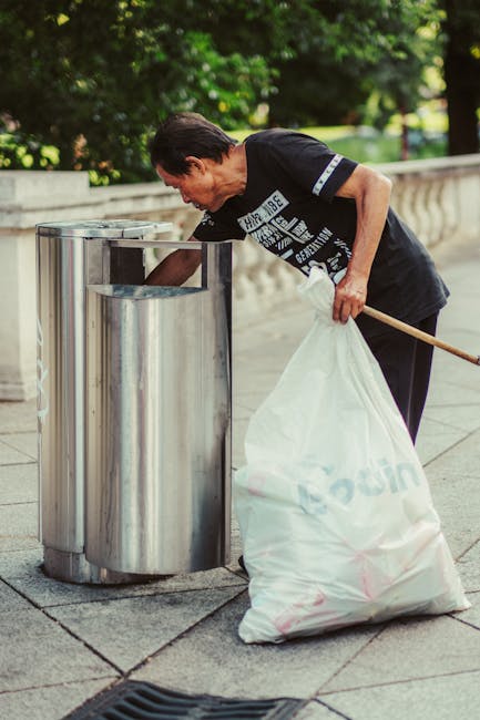 A woman with short dark hair, dressed in a black T-shirt and dark trousers, is seen disposing of rubbish into a modern, cylindrical stainless steel public trash bin located outdoors on a paved walkway. She is bending slightly forward, holding a large white plastic bag filled with waste in her left hand while using her right hand to open the bin's lid. The scene is set in an area with a decorative stone balustrade in the background, and lush green foliage and trees providing natural shade. The lighting appears natural, highlighting the reflective surface of the metal bin and the textured surface of the paving stones. This image relates to waste management and the importance of proper rubbish disposal, a key aspect of independent waste collection services provided by companies such as Waste Collection Catford, which offers alternative waste handling solutions outside of local authority schemes.