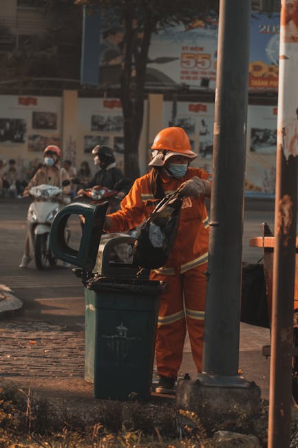 A sanitation worker dressed in an orange safety jacket, matching orange trousers, and a protective helmet, is seen collecting waste from a black trash bin placed on a sidewalk. The worker is wearing a face mask and gloves, and appears to be in the process of emptying or inspecting the bin, which is situated near a metallic pole that partially obstructs the view. In the background, there are several people riding scooters on the street, some wearing helmets and face masks, indicating adherence to safety protocols. Behind them, a wall with posters or advertisements is visible, along with trees and urban structures. The scene is illuminated by natural daylight, highlighting the textures of the worker’s uniform, the plastic surface of the trash bin, and the surrounding urban environment. This image reflects aspects of on-site waste collection by independent waste management services, such as Waste Collection Catford, operating within city streets and public spaces.