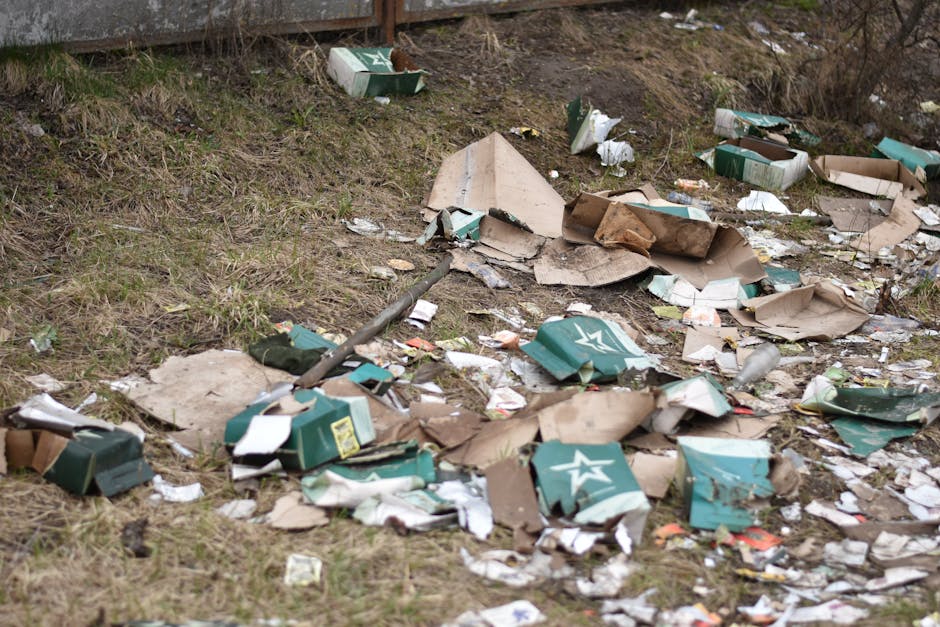A ground-level view of a patch of outdoor grass and soil, where piles of discarded cardboard boxes and packaging materials are scattered across the area. The cardboard boxes are predominantly green with white star logos, some flattened and crumpled, while others remain partially intact. There are also white and brown paper debris mixed among the packaging, and a few plastic bottles lying nearby. In the background, a wooden fence is visible, partially obscured by the litter, and some dried grass and small plants grow along the edge of the scene. The overall environment appears unkempt, indicating a location where illegal dumping or careless waste disposal has occurred, highlighting the type of debris often addressed by private waste removal services such as those offered by Waste Collection Catford. The lighting suggests daytime with overcast conditions, casting a neutral tone over the scene, emphasizing the need for outdoor rubbish clearance in residential or neglected areas.