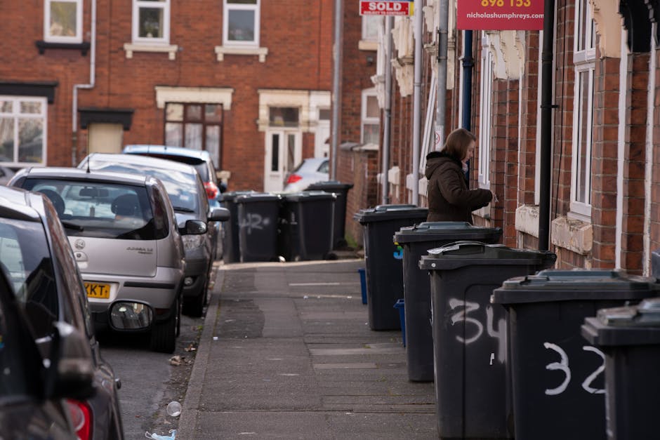 A residential street scene featuring a row of black wheelie bins with white numbering placed along the sidewalk in front of red brick terraced houses. The bins are positioned close to the buildings' front walls, some with partially open lids. A woman wearing a dark coat stands next to the nearest bin on the right, appearing to be managing rubbish or waste. The sidewalk is made of concrete slabs, with a narrow strip of asphalt road on the left side, where several parked cars are aligned facing away from the camera. The vehicles vary in color and model, including a silver hatchback and dark-colored cars. The background shows more houses with similar architectural styles, large windows with white frames, and some windows partially covered by blinds or curtains. Above the scene, a red sign with white and yellow text is attached to the wall, suggesting an estate or property sale, with a website address included. The overall atmosphere indicates an area where private waste collection services, such as waste collection Catford, offer an alternative to local authority rubbish removal, with a focus on curbside waste management and on-site disposal.