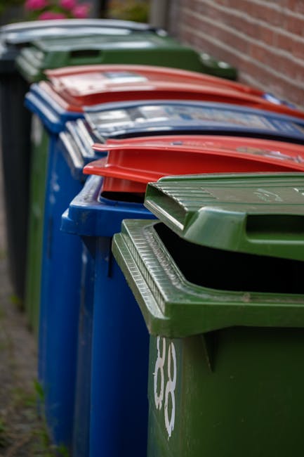 A row of large wheelie bins lined up along a paved sidewalk beside a brick wall, with the earliest bin in the foreground being dark green with a closed lid, followed by a blue bin with a closed lid, then a red bin and a further dark green bin, all with textured, ridged lids. The bins are positioned on the edge of the pavement, and the background shows a blurred glimpse of additional bins and the street environment. The scene depicts standard waste collection containers, which are typically used for household rubbish disposal in private waste management services. The image captures the utilitarian appearance of these bins, emphasizing their metallic construction and practical design. The natural daylight highlights the different colours and textures of the lids and sides, reflecting typical outdoor waste storage. This arrangement of bins aligns with the context of private rubbish collection and alternative waste handling, often seen outside residential properties before scheduled collection or during on-site clearance activities, as provided by waste collection services such as Waste Collection Catford.