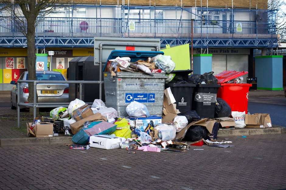 A collection of overflowing rubbish bins and surrounding waste on a paved urban street, including large black and black mixed-paper-and-cardboard recycling bins, a red bin, and a tan recycling box. The waste, which comprises various flattened cardboard boxes, plastic bags, paper, and other miscellaneous refuse, is piled around the bins and spilling onto the pavement. Behind the waste collection area, a metal railing separates the bins from a parked silver car with a visible yellow registration plate. The background features a storefront with signs and a blue construction fence, indicating nearby construction work. The scene is lit naturally, suggesting daylight, and reflects an instance of non-collection or excess waste accumulated outside a designated rubbish collection point, characteristic of private waste handling or alternative rubbish removal services like those offered by Waste Collection Catford in a commercial or residential locality on Catford Broadway.